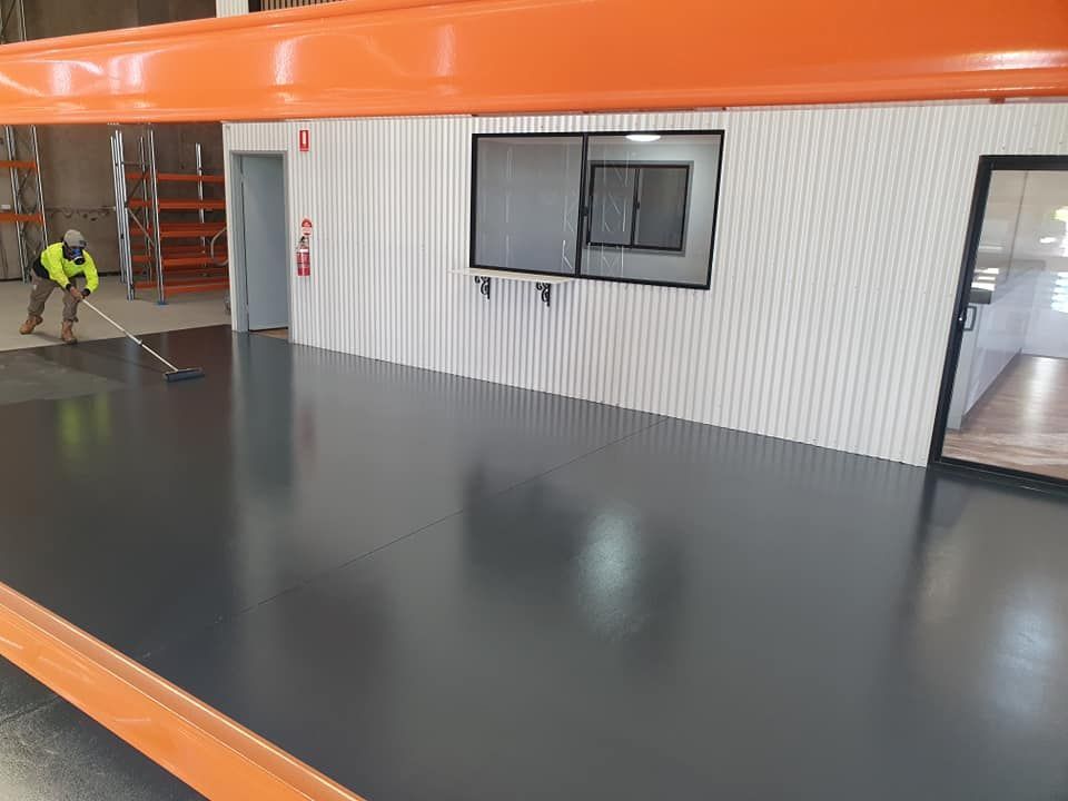 A Man Is Painting The Floor Of A Warehouse With A Broom — LMS Painting & Maintenance Pty Ltd In North Lambton, NSW