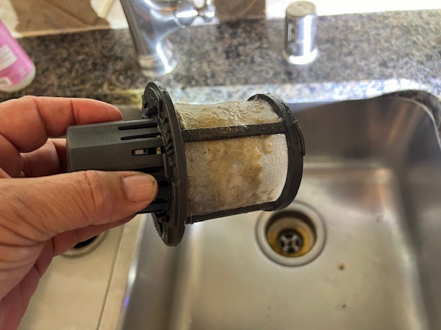 Hand holding a dirty dishwasher filter over a kitchen sink. The filter is covered in food residue.