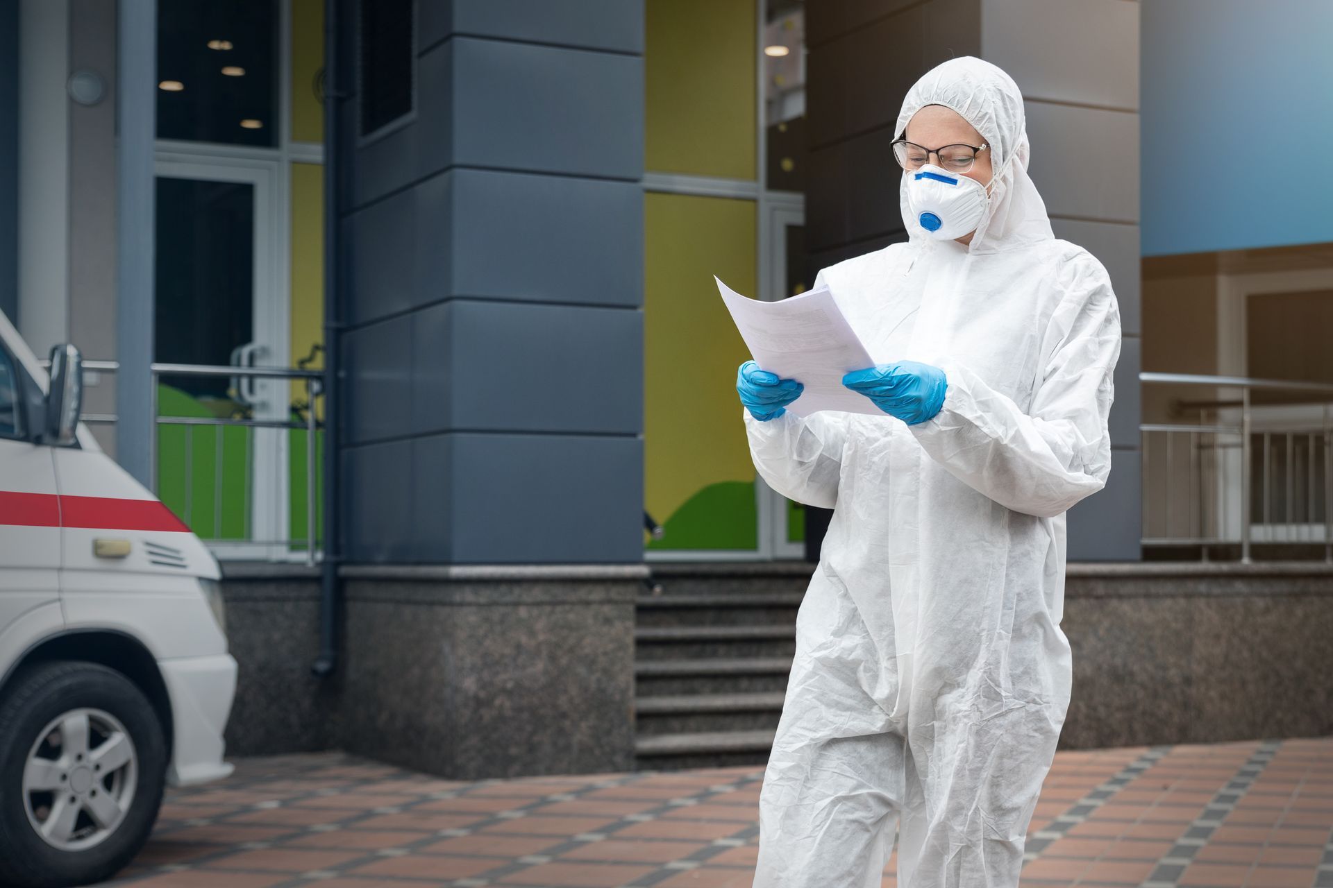 a close up of a wall with a wooden frame Person in white hazmat suit, mask, and gloves reads papers near ambulance and building entrance.