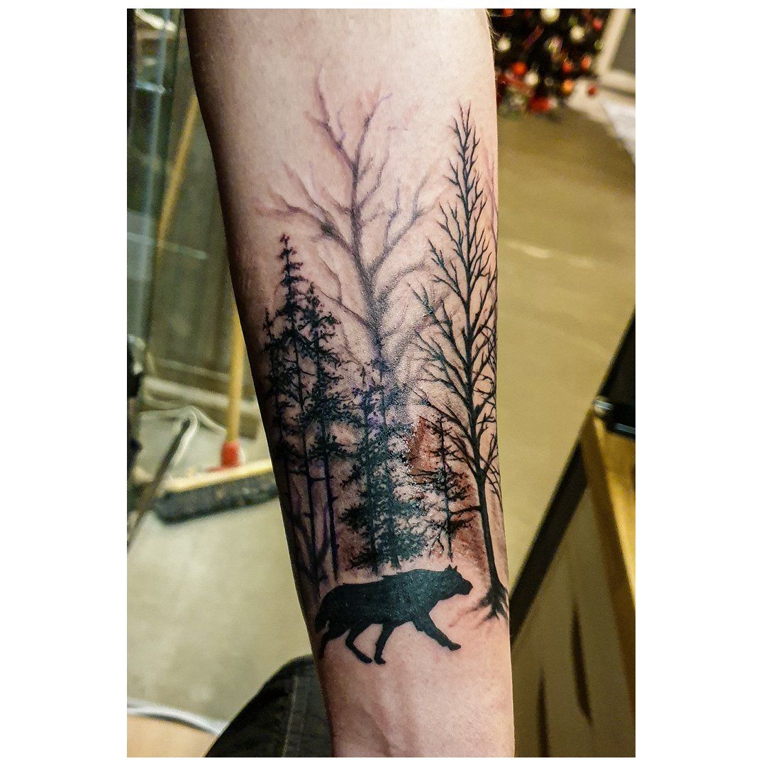 Een tatoeage van een wolf die door een bos loopt op iemands onderarm.