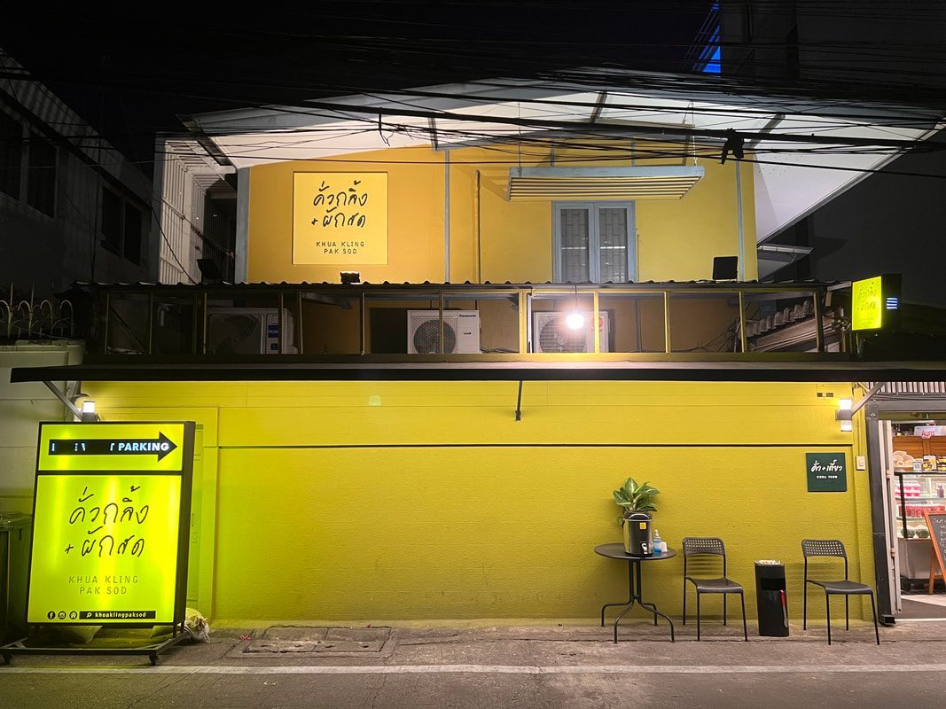 Yellow building with a sign in Thai, at night, with a table and chairs set up outside.
