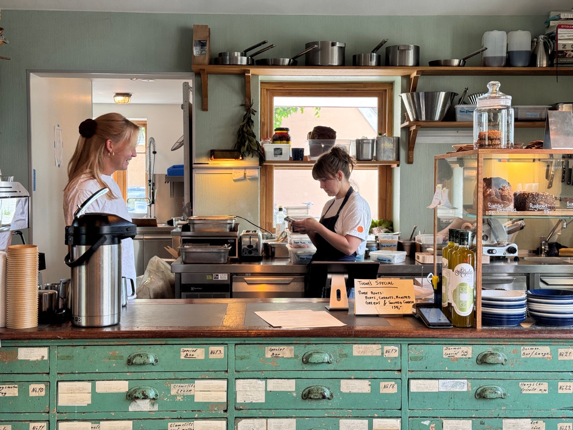 Restaurant exterior with two men seated, motorbike parked. Pink awning, white building.
