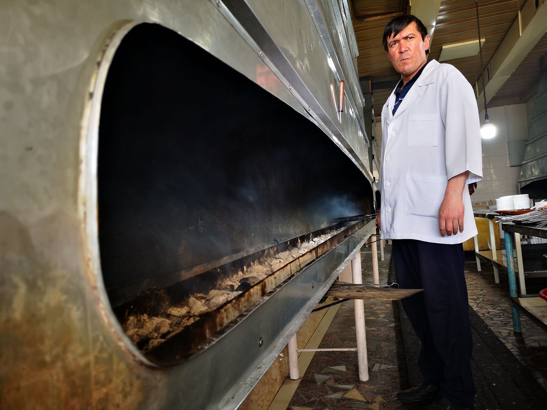 Man in white coat stands next to a large oven, possibly for cooking meat.