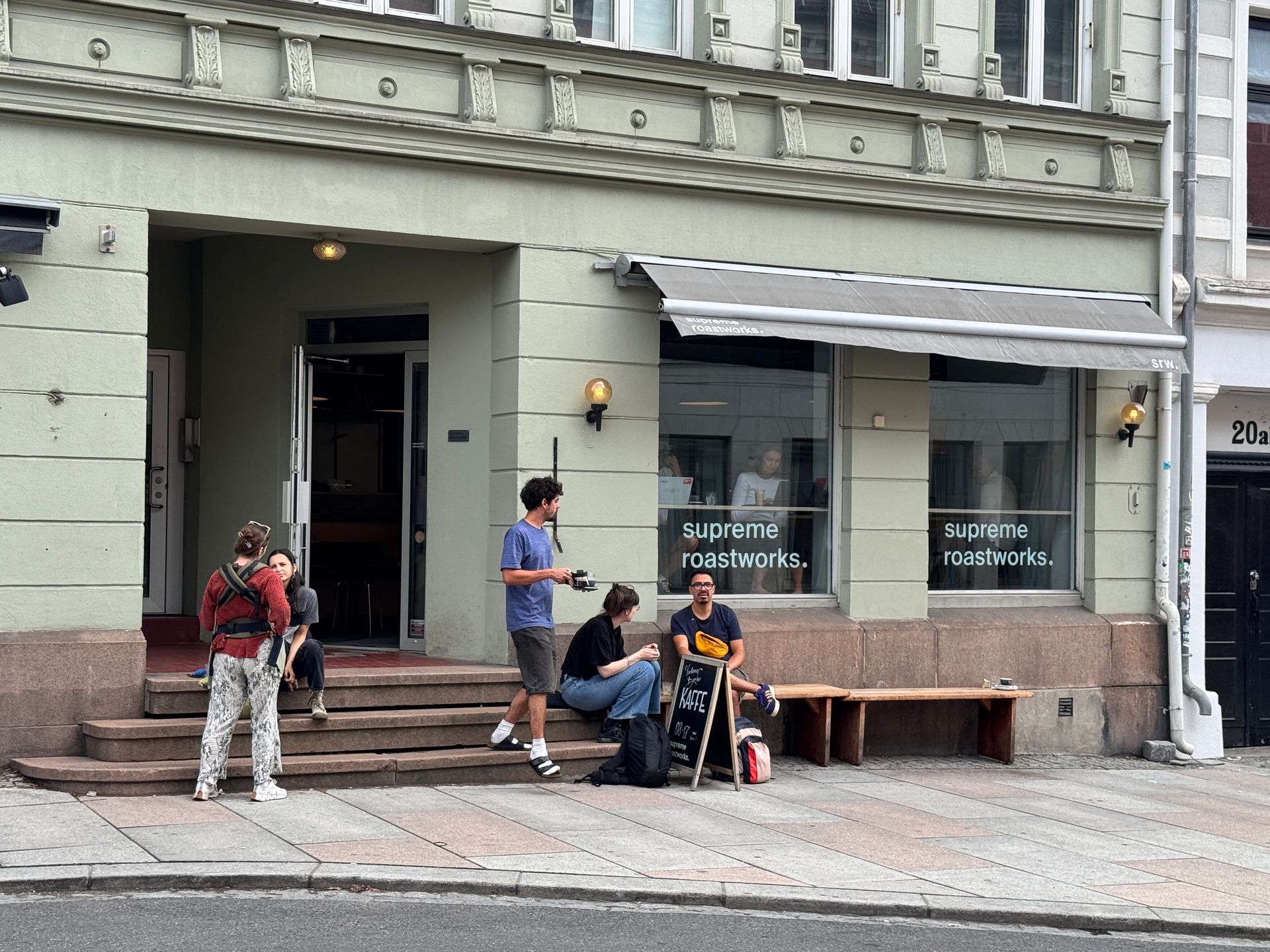 Restaurant exterior with two men seated, motorbike parked.  Pink awning, white building.