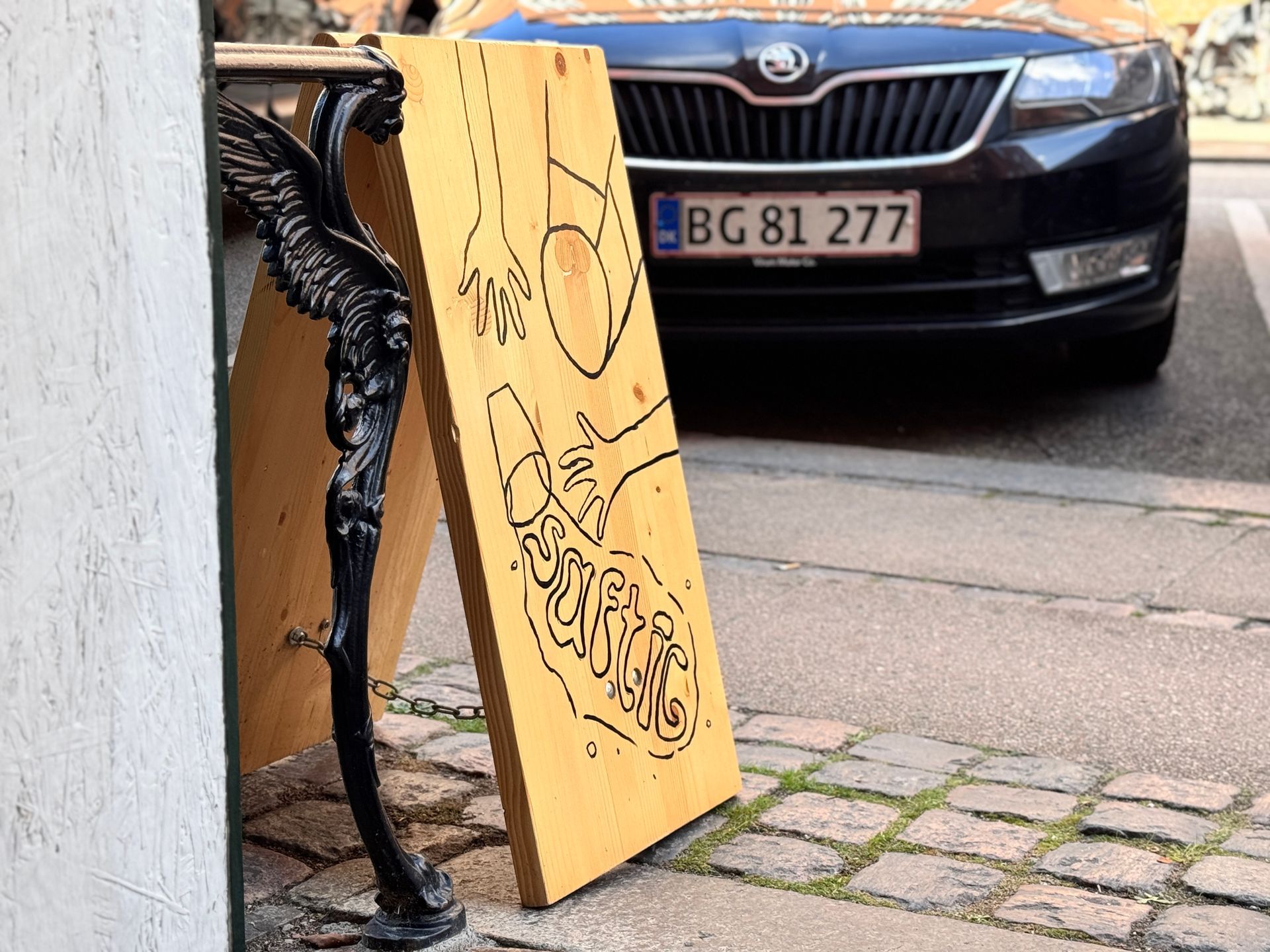 Wooden sign with hand-drawn images leaning on a decorative iron post. A car with license plate BG 81 277 is in the background.