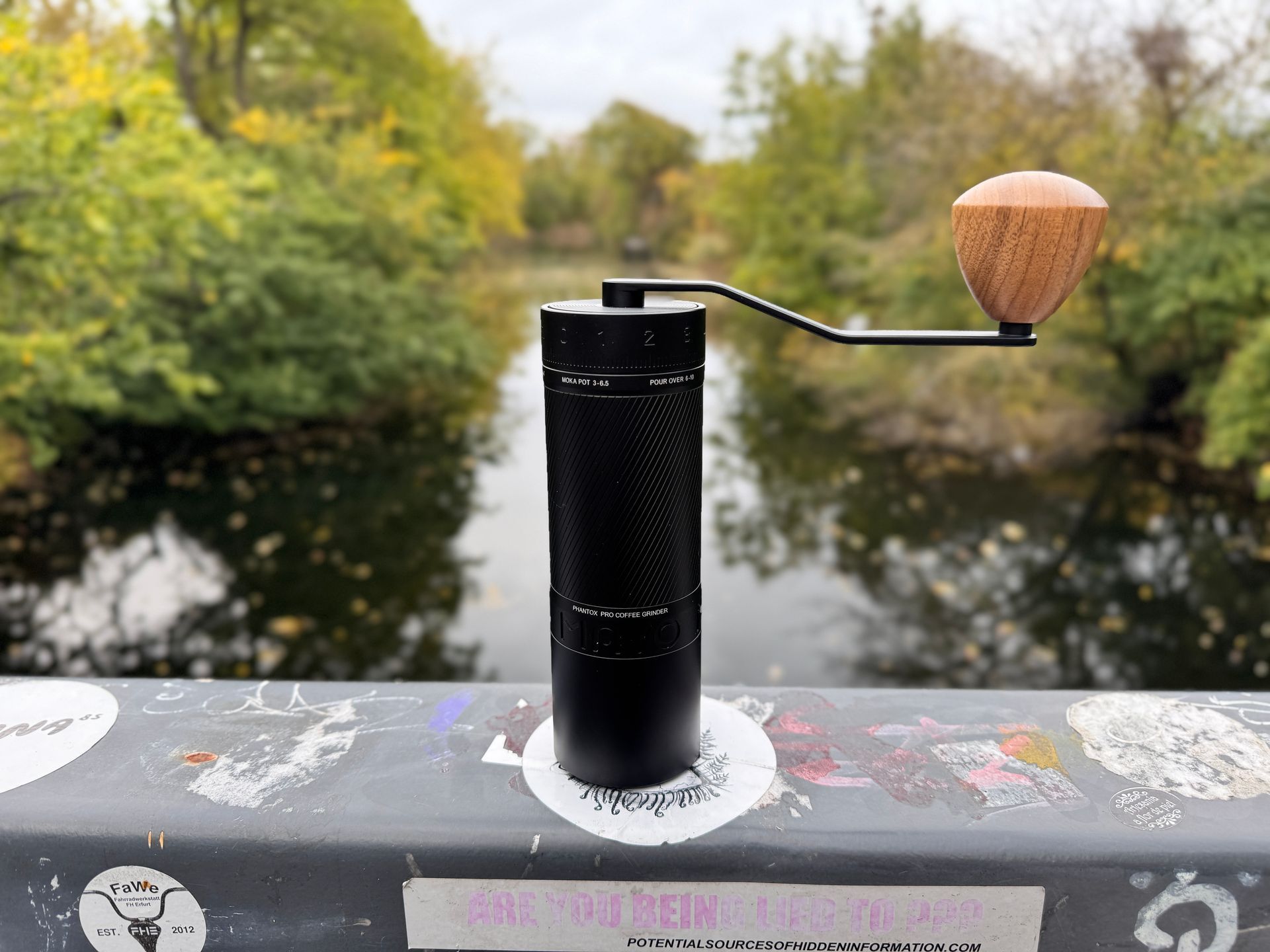 Black hand coffee grinder on a bridge railing, with river and foliage in the background.