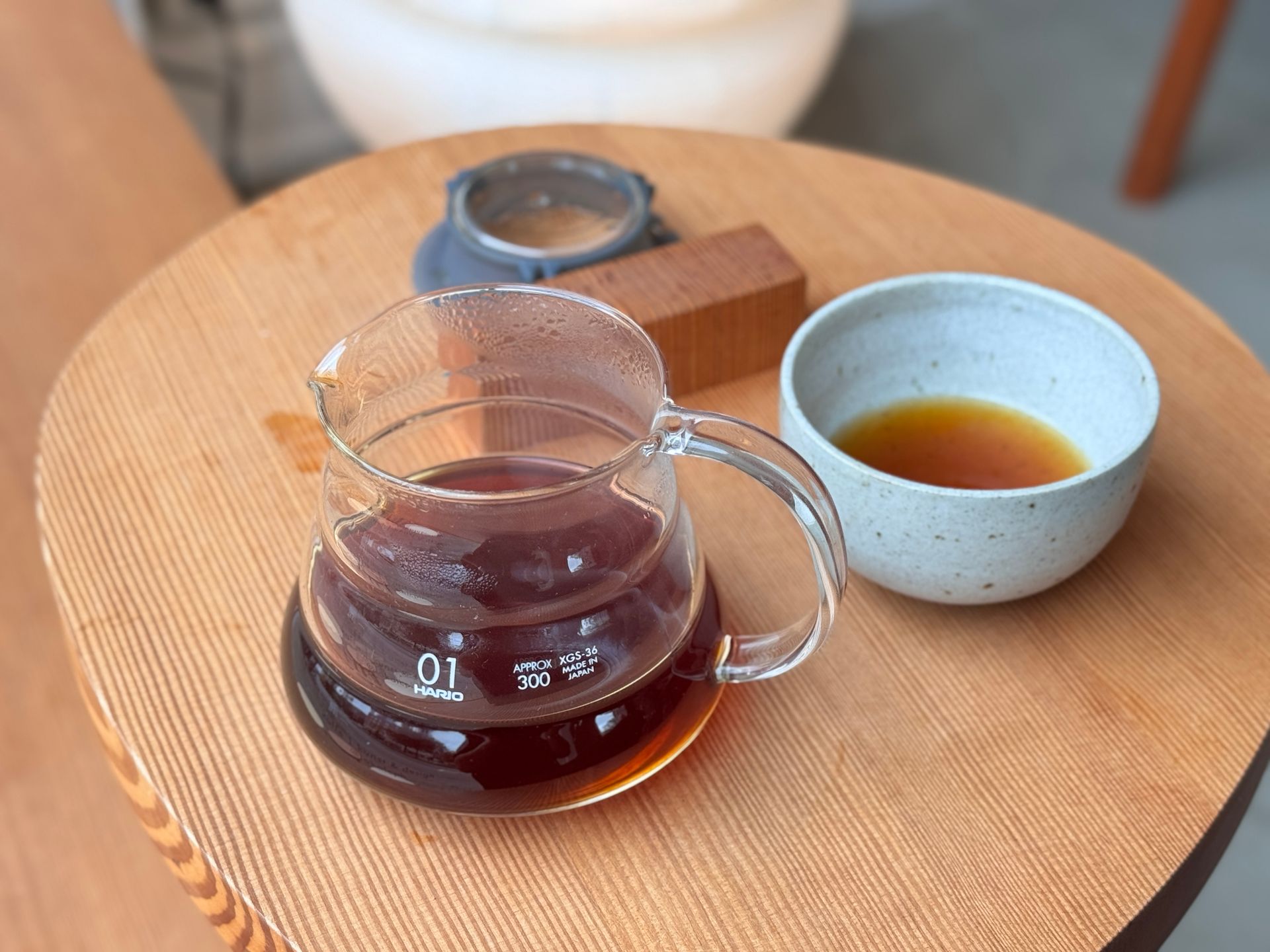 Glass carafe of coffee, a small bowl of coffee, and filter equipment on a wooden table.
