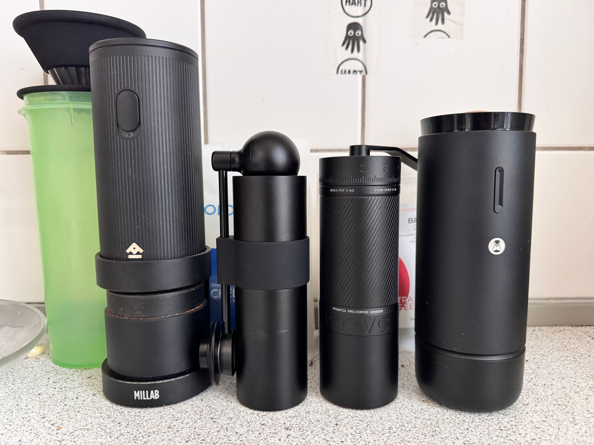 Four black coffee grinders of different designs on a counter, with a green container on the left.