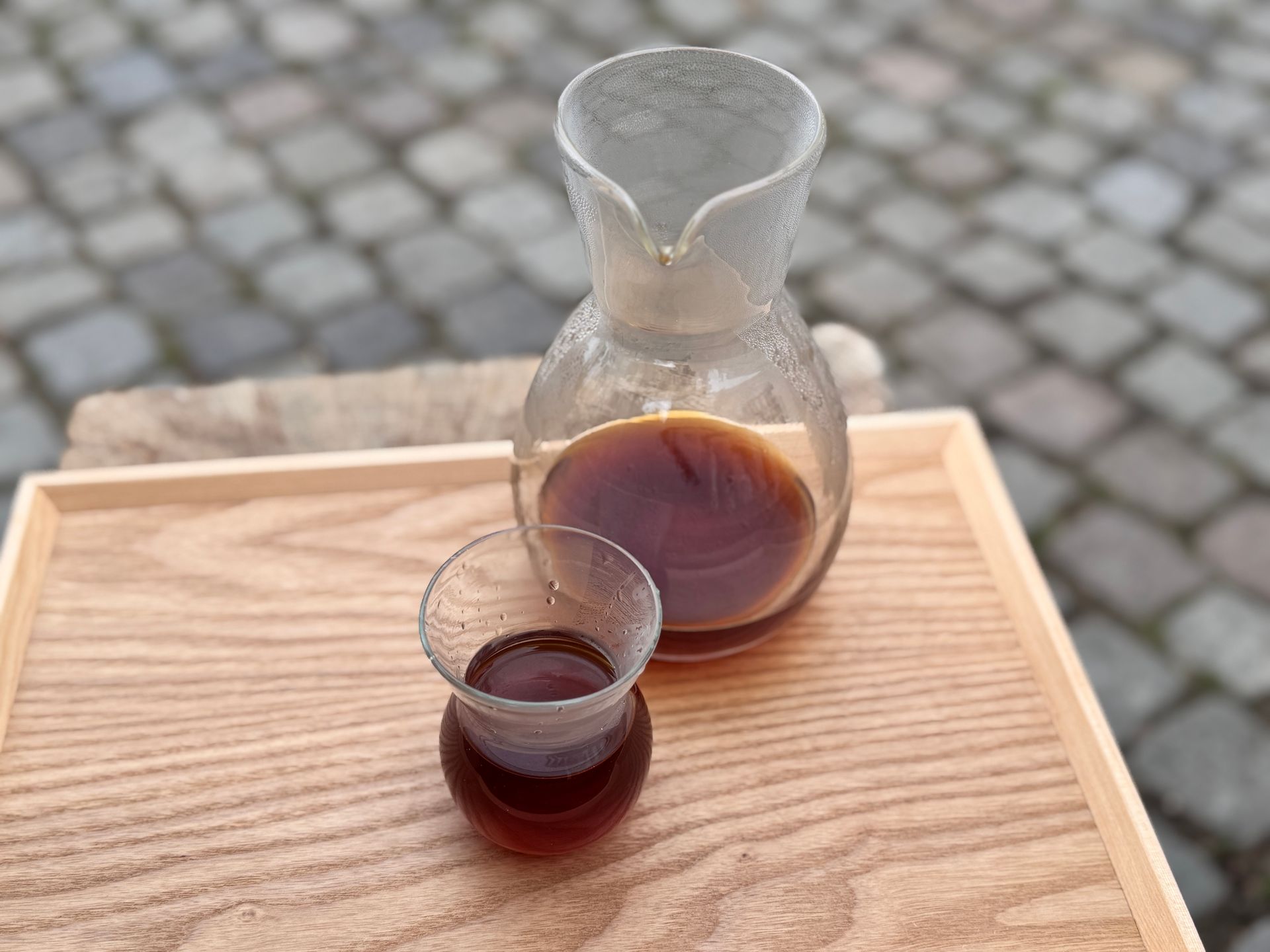 Coffee, glass carafe, and small glass on wooden tray, set on cobblestone.