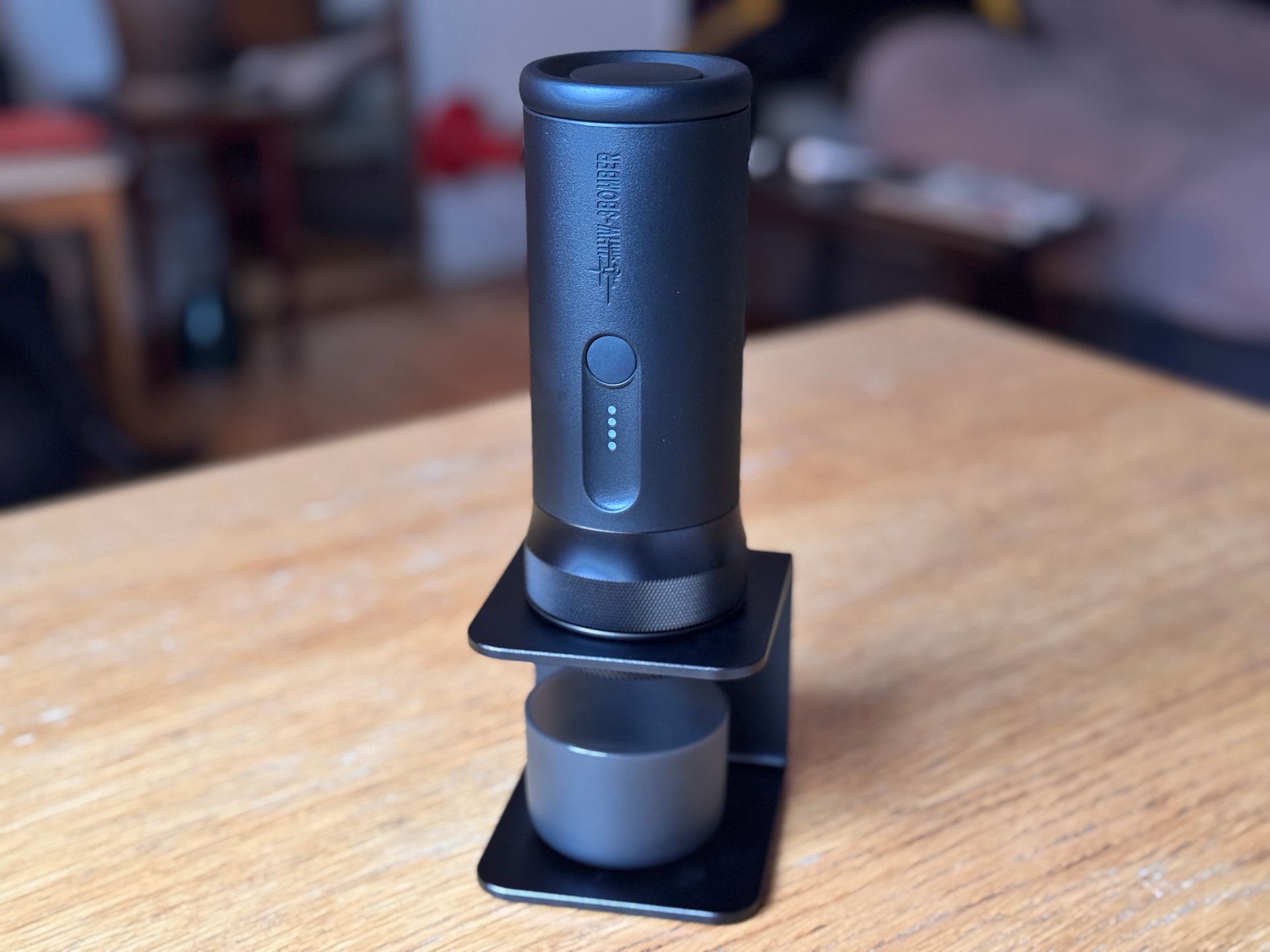 Black manual espresso maker on a stand, placed on a wooden table. Blurred background.