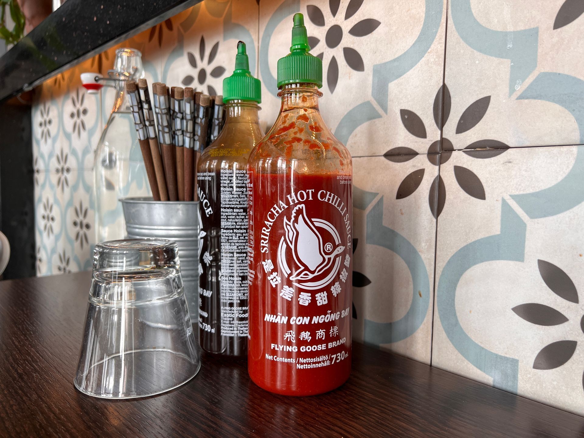 Bottles of sriracha and chili sauce on a wooden counter in front of decorative tile.