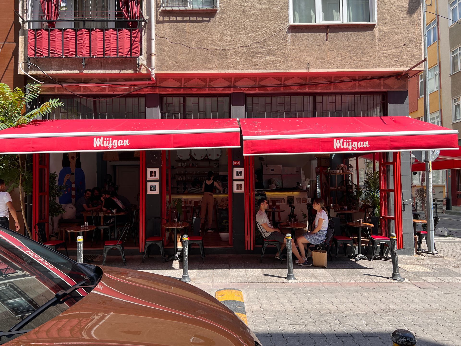 Restaurant exterior with two men seated, motorbike parked.  Pink awning, white building.