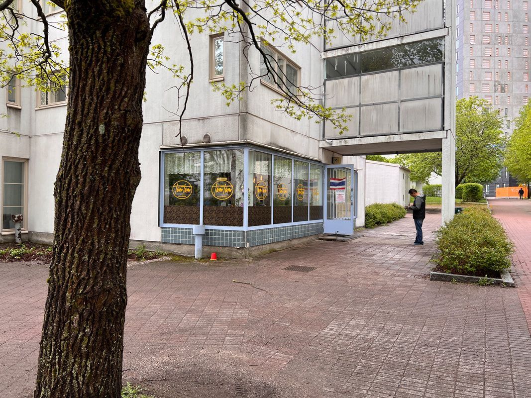 Restaurant exterior with two men seated, motorbike parked.  Pink awning, white building.