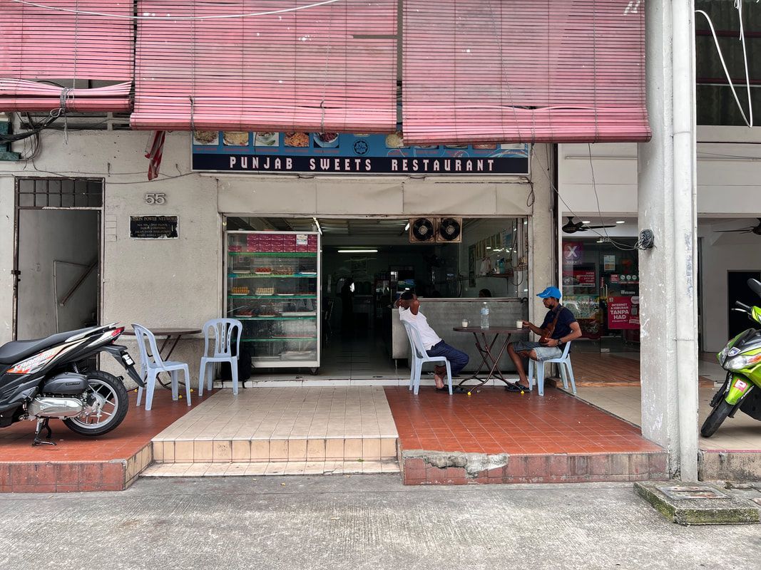Restaurant exterior with two men seated, motorbike parked. Pink awning, white building.