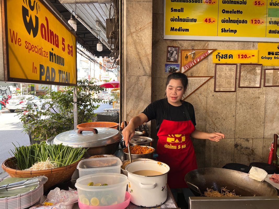 Woman in red apron cooks street food at a stall. Vegetables and ingredients on the counter.