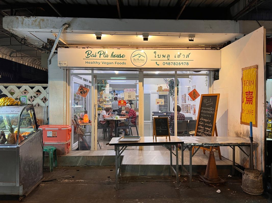 A small restaurant with a white facade, glass door, and outdoor tables. A yellow sign hangs on the right.