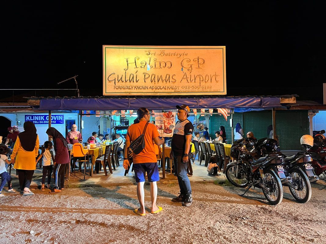 People at a night market restaurant, motorcycles parked nearby. Sign reads: Bakar Gulai Panas Airport