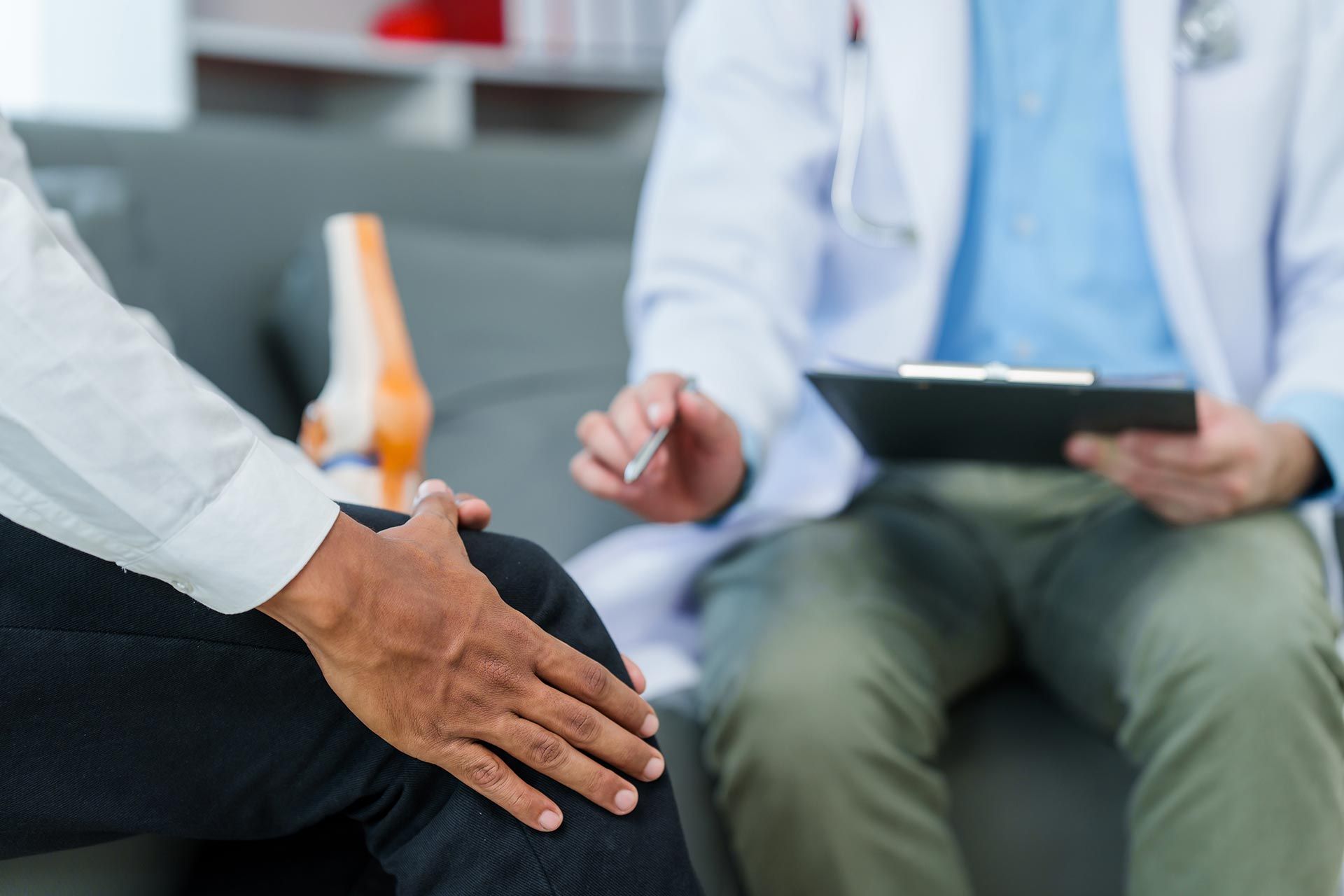 Patient seated during a medical consultation while a clinician holds a clipboard and takes notes.