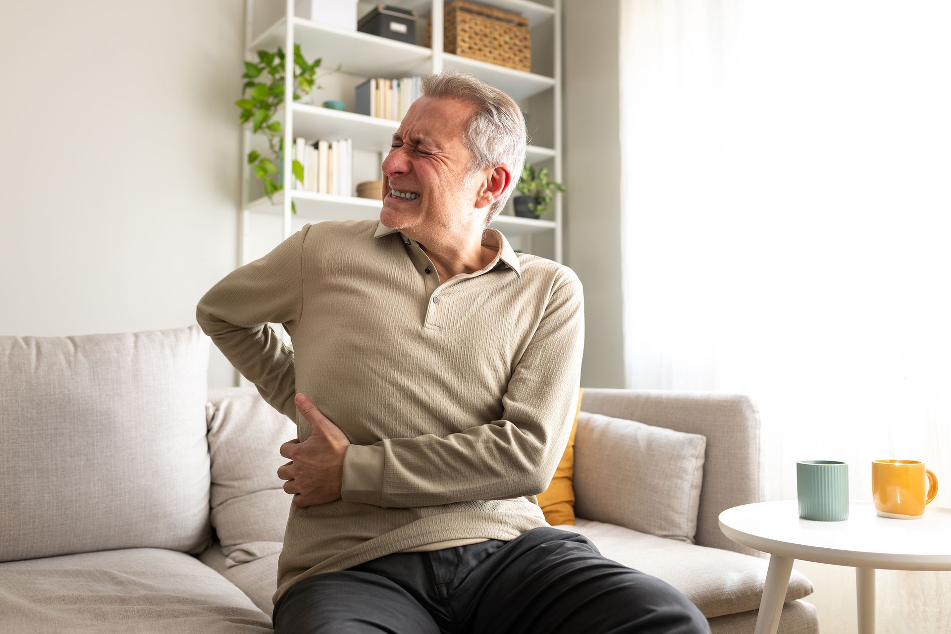 Older male adult on couch clutches back in pain near bright window.