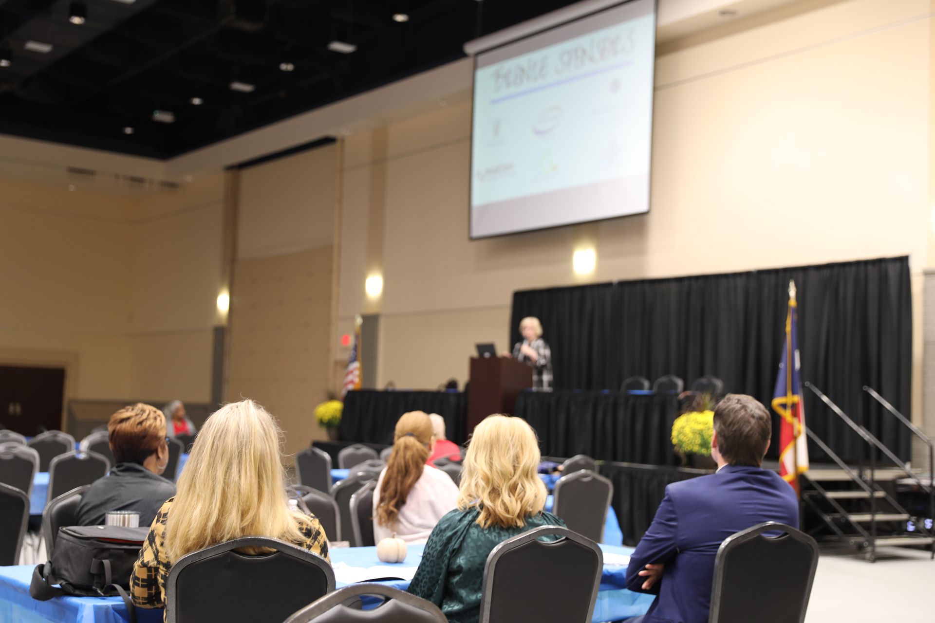 A group of people are sitting at tables in a conference room watching a presentation.