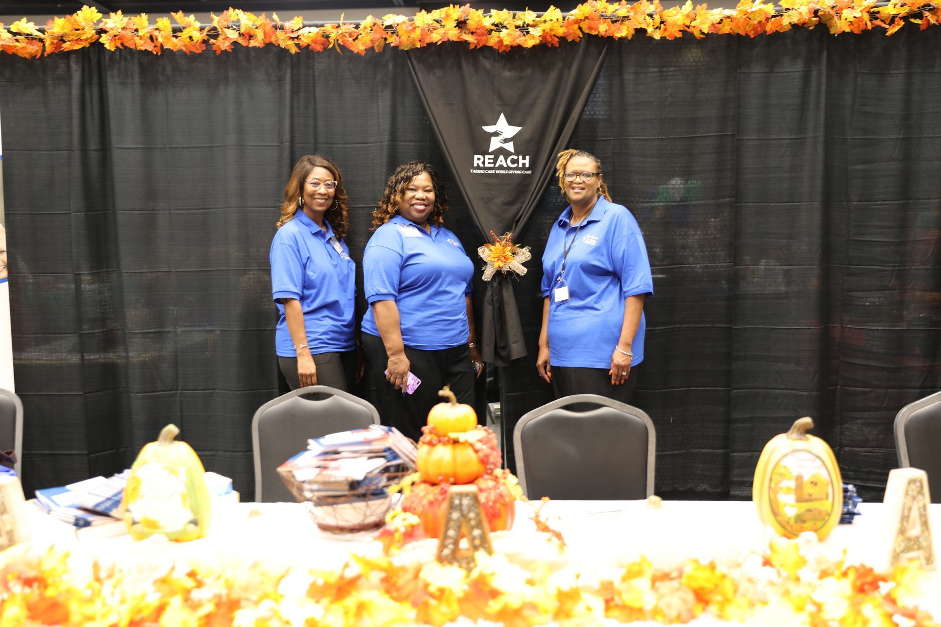 Three women in blue shirts are standing in front of a table with pumpkins and leaves on it.