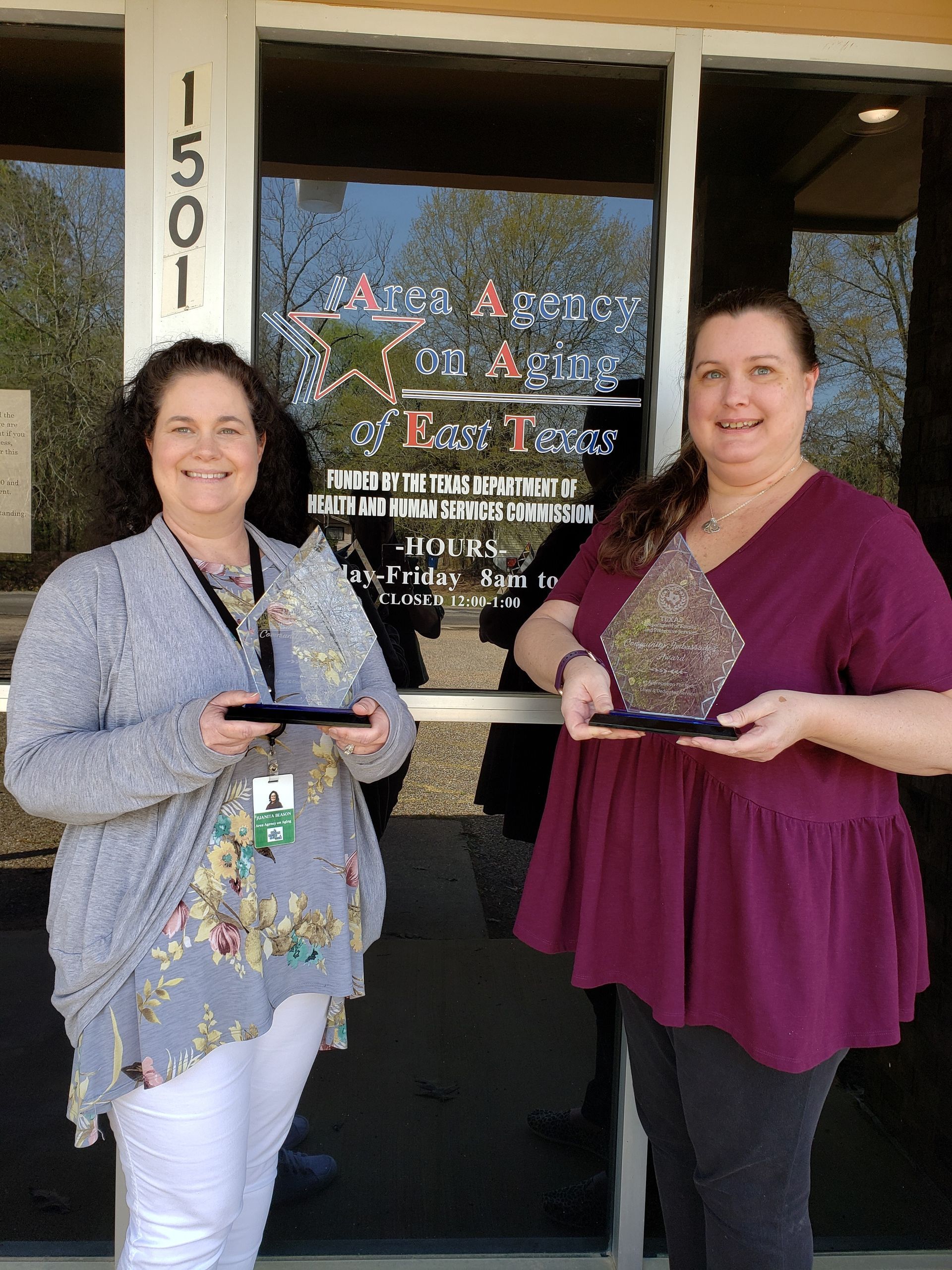 Two women are standing in front of a building holding trophies.