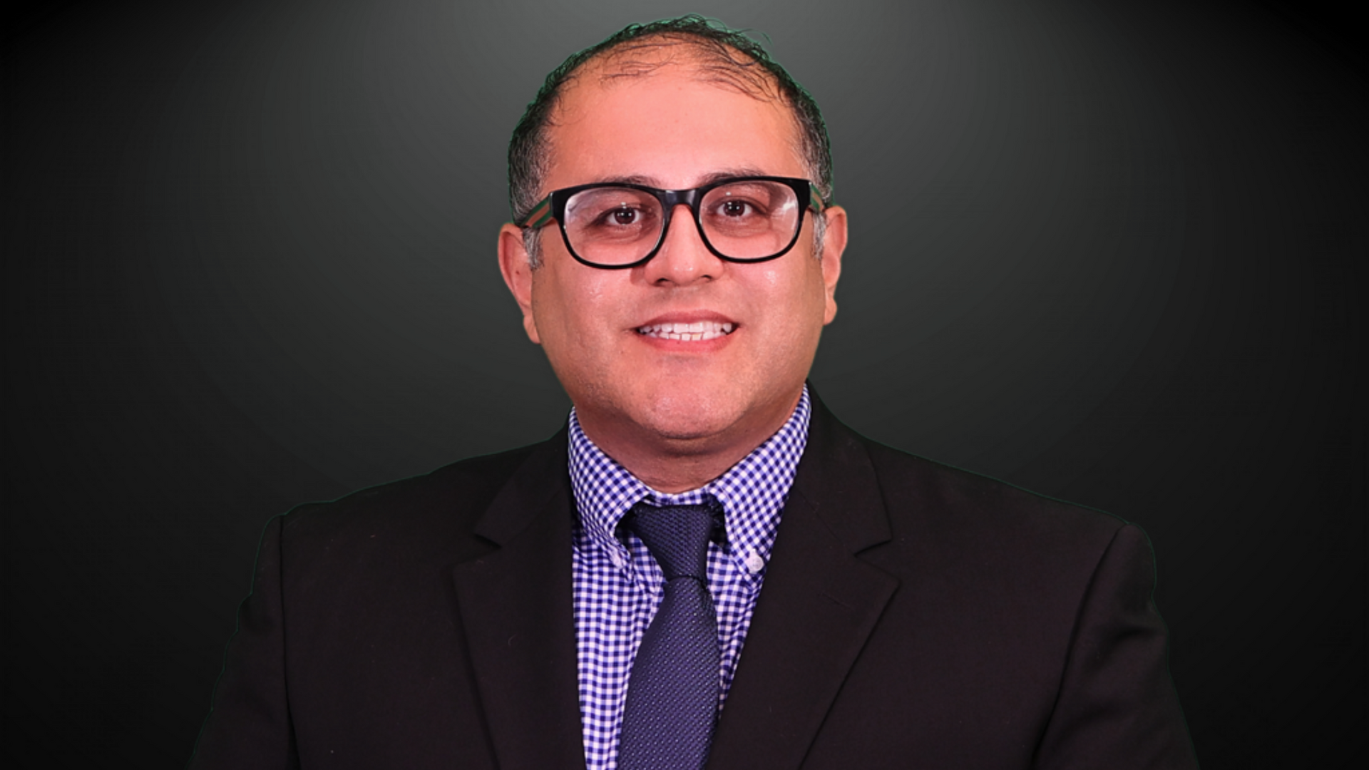 Man in glasses and suit smiles at the camera against a dark background.