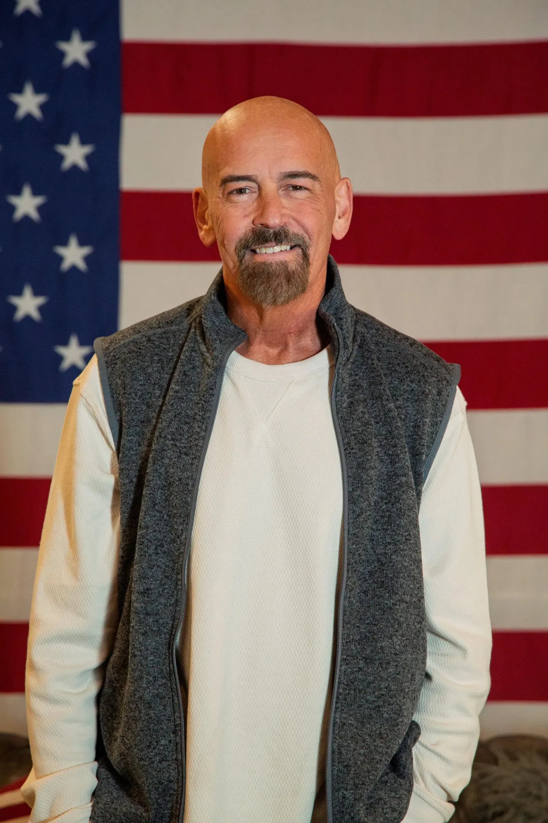 John Deaton is image shows a man with a beard and shaved head smiles standing in front of an American flag.