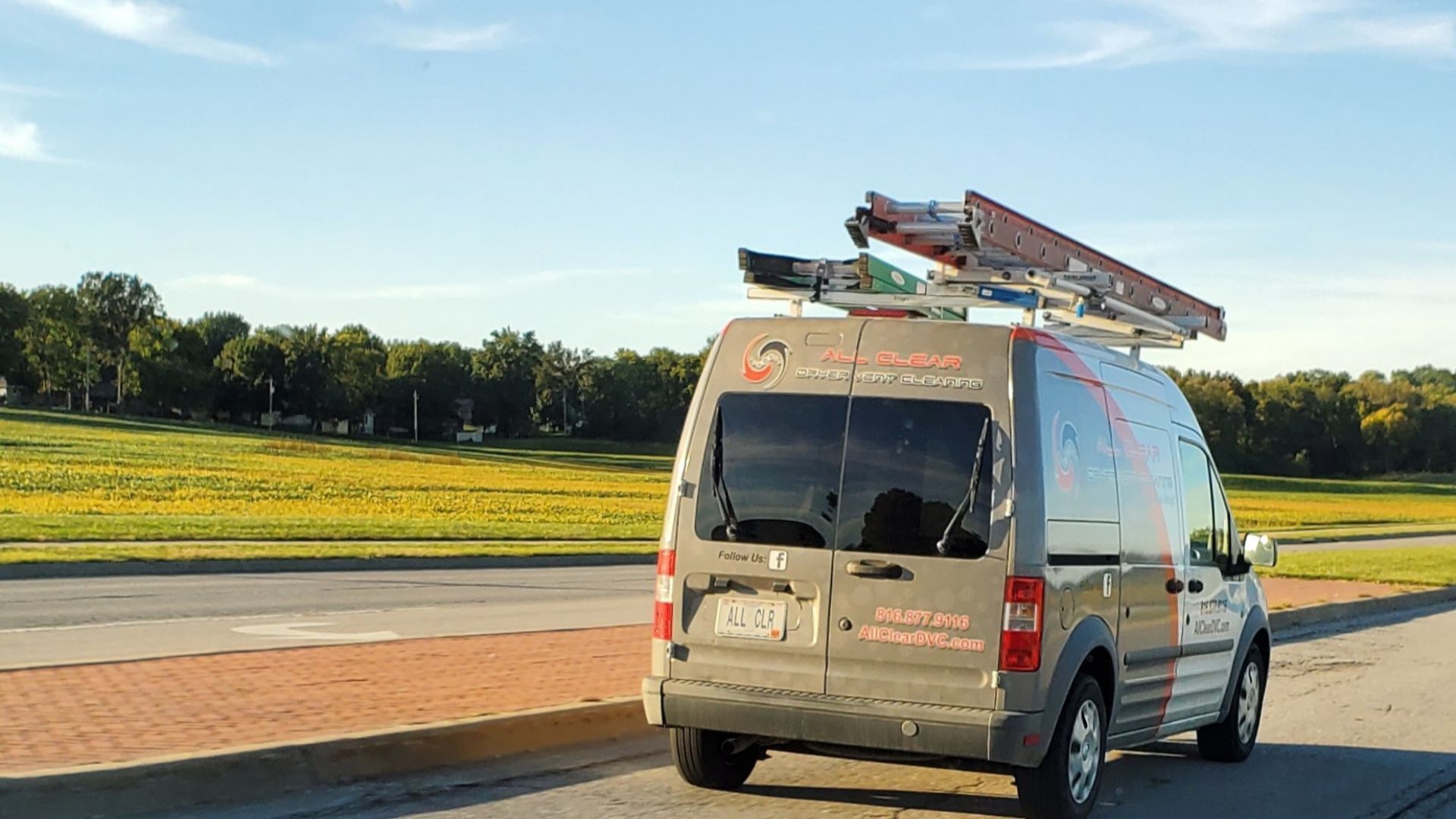 Silver utility van driving on a road beside a grassy field, with ladders strapped to the roof.