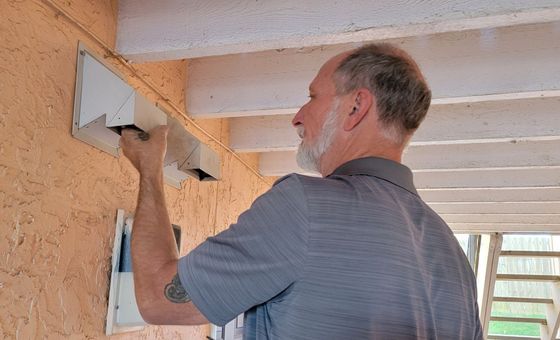 Man installing a white metal bracket on an orange stucco wall outdoors under a porch ceiling