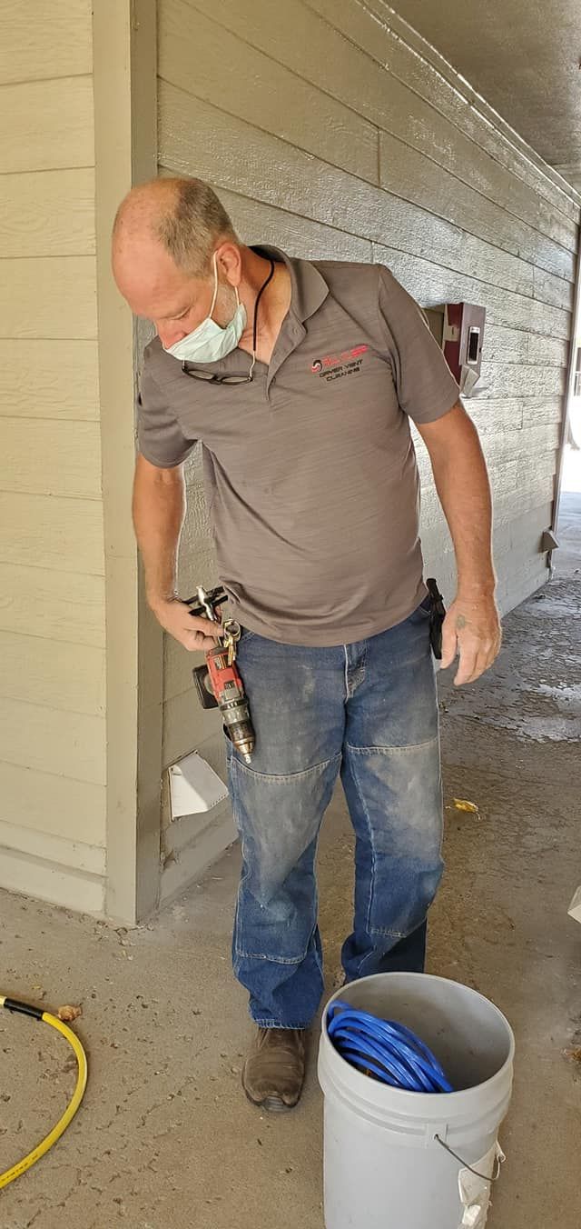 Worker in a gray shirt sanding a wall inside a room, with a white bucket and hose on the floor.