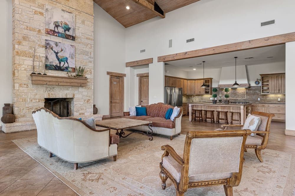Spacious living room with high ceilings and exposed wooden beams, featuring a stone fireplace adorned with two abstract paintings of deer. Elegant wooden furniture, including a sofa, two armchairs, and a rustic coffee table, fill the space. A large area rug covers the tiled floor. The open-concept design leads into a kitchen area with wooden cabinetry and a central island with bar stools at Marquis at Canyon Ridge in Austin, TX.