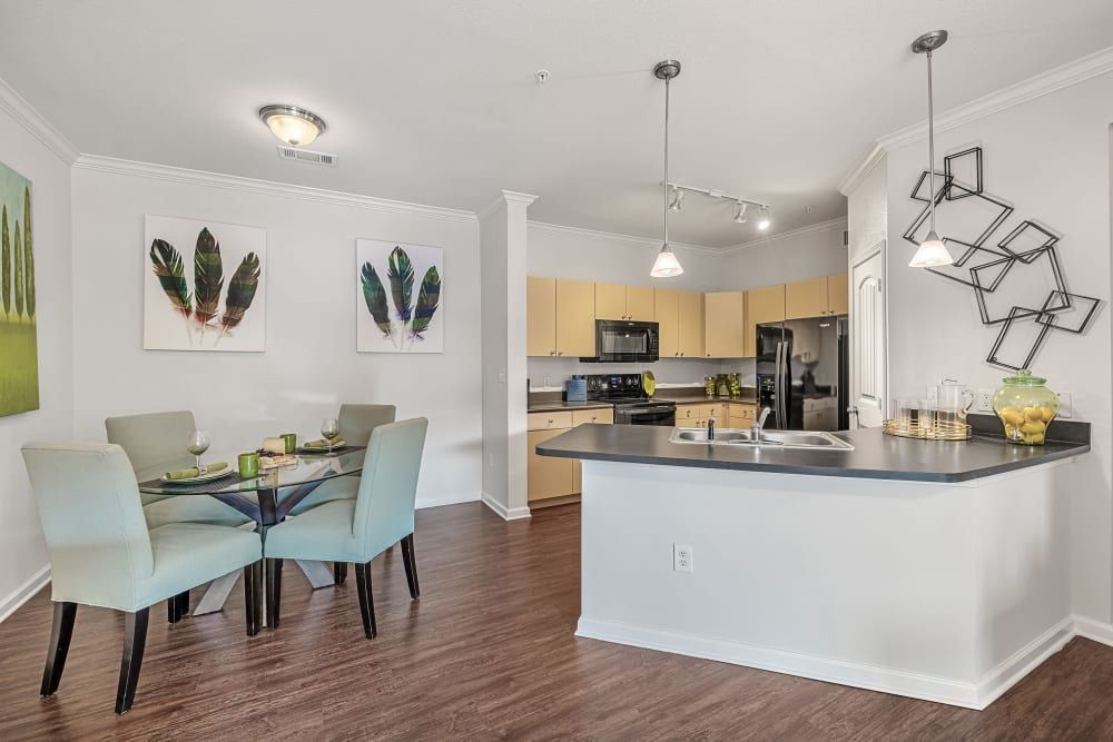 Modern, open-concept apartment interior featuring a dining area with a glass-top table and mint green chairs, with decorative feather artwork on the walls. The adjoining kitchen has beige cabinets, black appliances, and a breakfast bar with a geometric metal wall sculpture above it. The space is well-lit with both pendant and ceiling lights, complementing the dark wood flooring at Marquis at Canyon Ridge in Austin, TX.