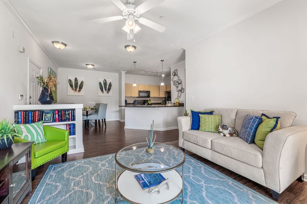 Brightly lit open-plan living room with a beige sofa adorned with blue and green pillows, a cozy dog resting on the couch, a matching blue and white patterned rug on the floor, and a clear glass coffee table with a book on it. On the left, there's a green armchair alongside a bookshelf filled with books and potted plants. In the background is a dining area with a dark wood table and chairs, leading to a kitchen with modern appliances and pendant lighting. The walls are decorated with framed leaf prints, and the space has a warm and inviting feel at Marquis at Canyon Ridge in Austin, TX.