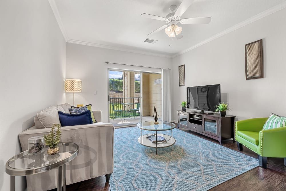 Modern living room interior with natural light, featuring a beige sofa with blue and beige cushions, a bright green accent chair with a striped pillow, a blue patterned area rug, glass-top coffee and side tables, a television on a wooden media console, white walls, hardwood floors, and a ceiling fan. A sliding glass door opens to a balcony with a view of a fence and greenery at Marquis at Canyon Ridge in Austin, TX.