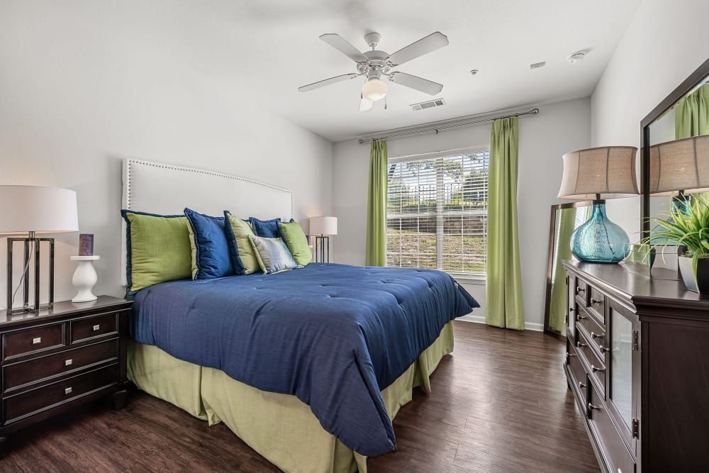 A modern bedroom with a large bed covered in a navy-blue comforter and an assortment of green and blue pillows. The bed has a white tufted headboard and is flanked by two dark wood nightstands with matching lamps. A dresser with a mirror above it is against the wall opposite the bed, next to a window dressed with sheer green curtains. There's a ceiling fan above, and the room has hardwood floors. Decorative items, including a turquoise vase and green plants, add pops of color at Marquis at Canyon Ridge in Austin, TX.