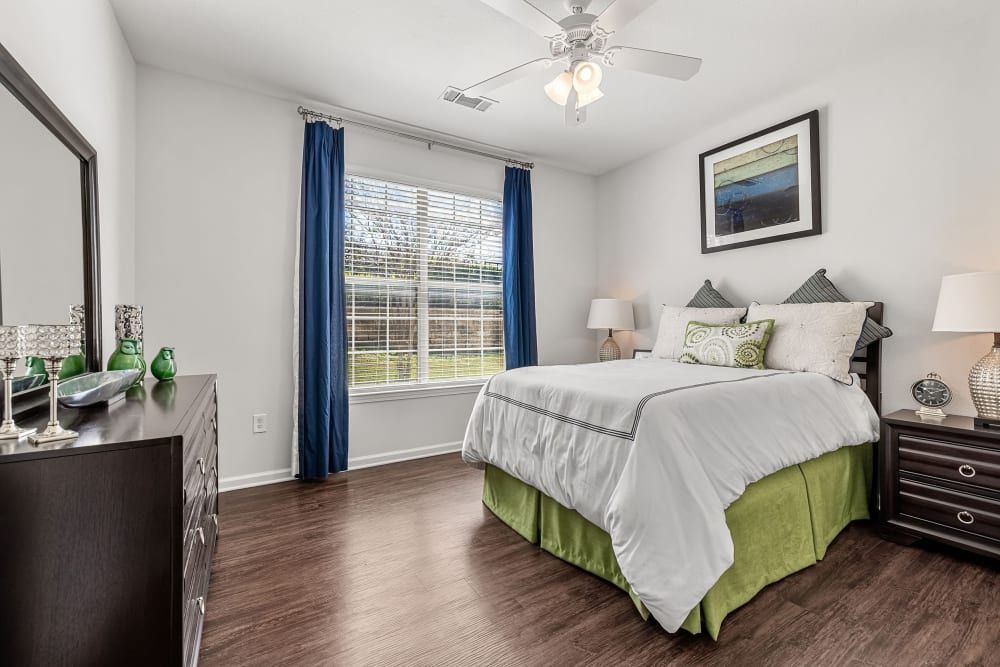 A modern bedroom with a large window draped in blue curtains. The bed is covered with white and green bedding and flanked by dark wooden nightstands with table lamps. Above the bed, there's a framed landscape painting, and to the left, a dresser topped with decorative items is visible. The room has hardwood floors and a white ceiling fan at Marquis at Canyon Ridge in Austin, TX.