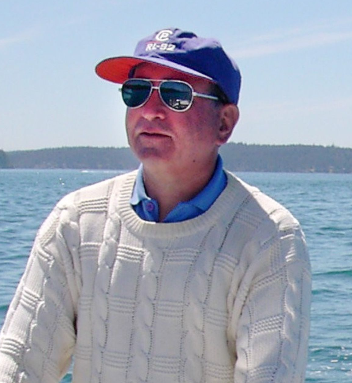 A person wearing a blue cap, sunglasses, and a cream cable-knit sweater standing in front of a lake.
