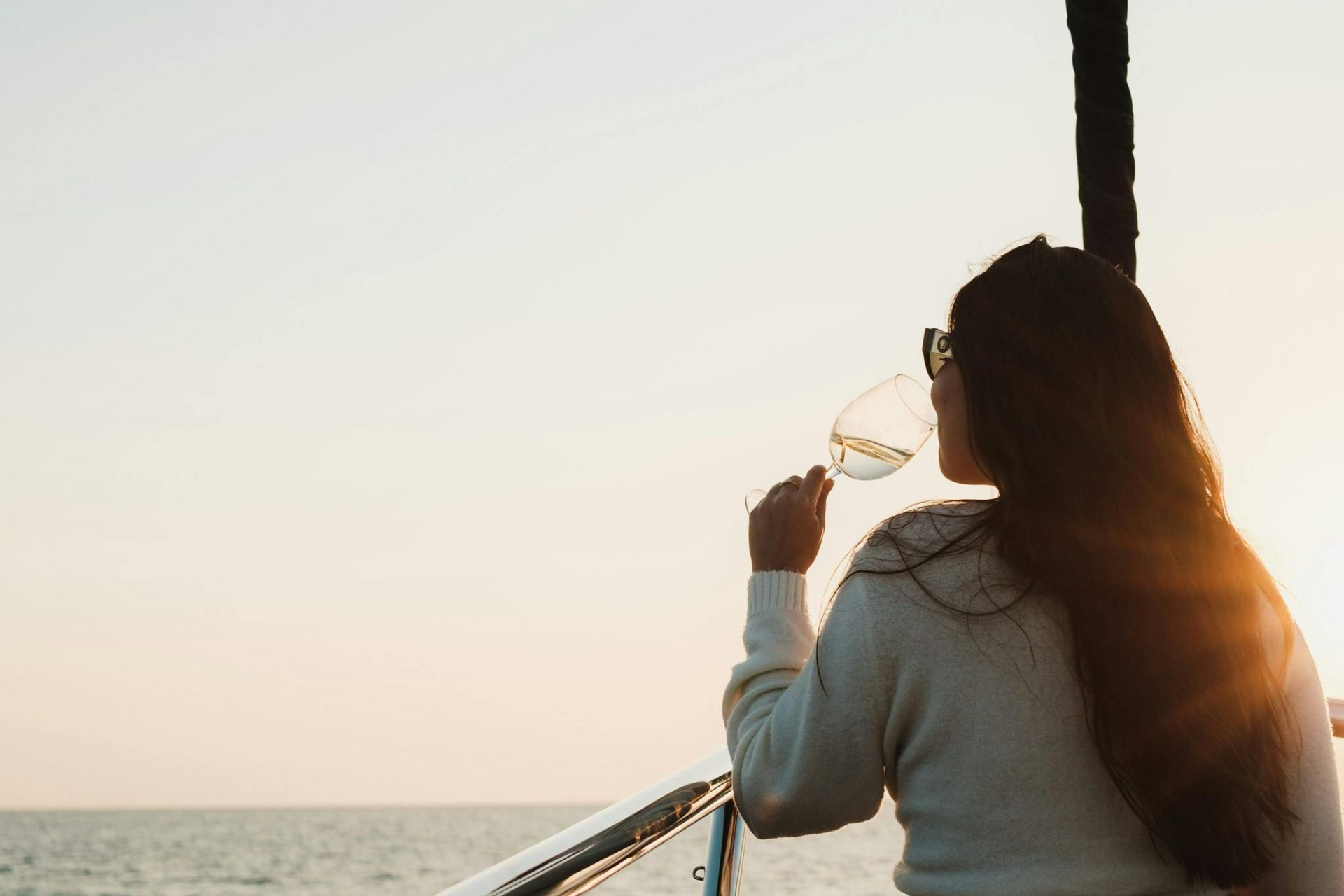 A person with long dark hair, wearing a white sweater, holds a wine glass while looking out at the ocean during sunset.