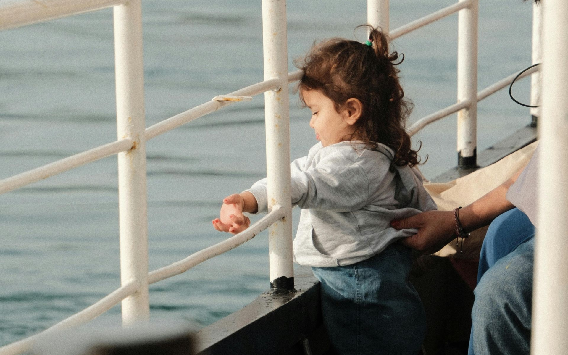 A toddler in a gray sweatshirt and blue pants reaches through a white metal railing toward water on a boat deck.