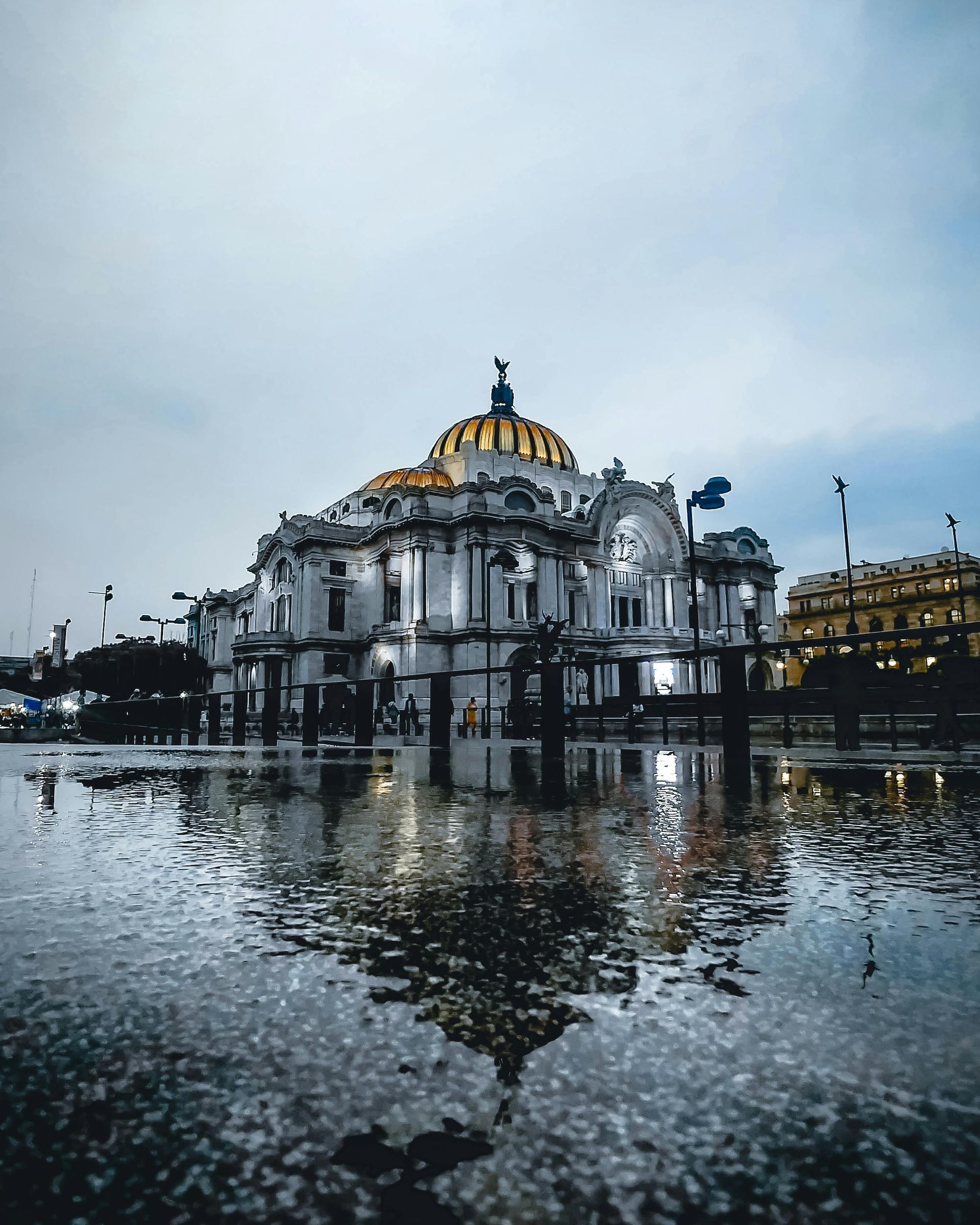 The Palacio de Bellas Artes in Mexico City at dusk, reflected in a large rain puddle on the ground.