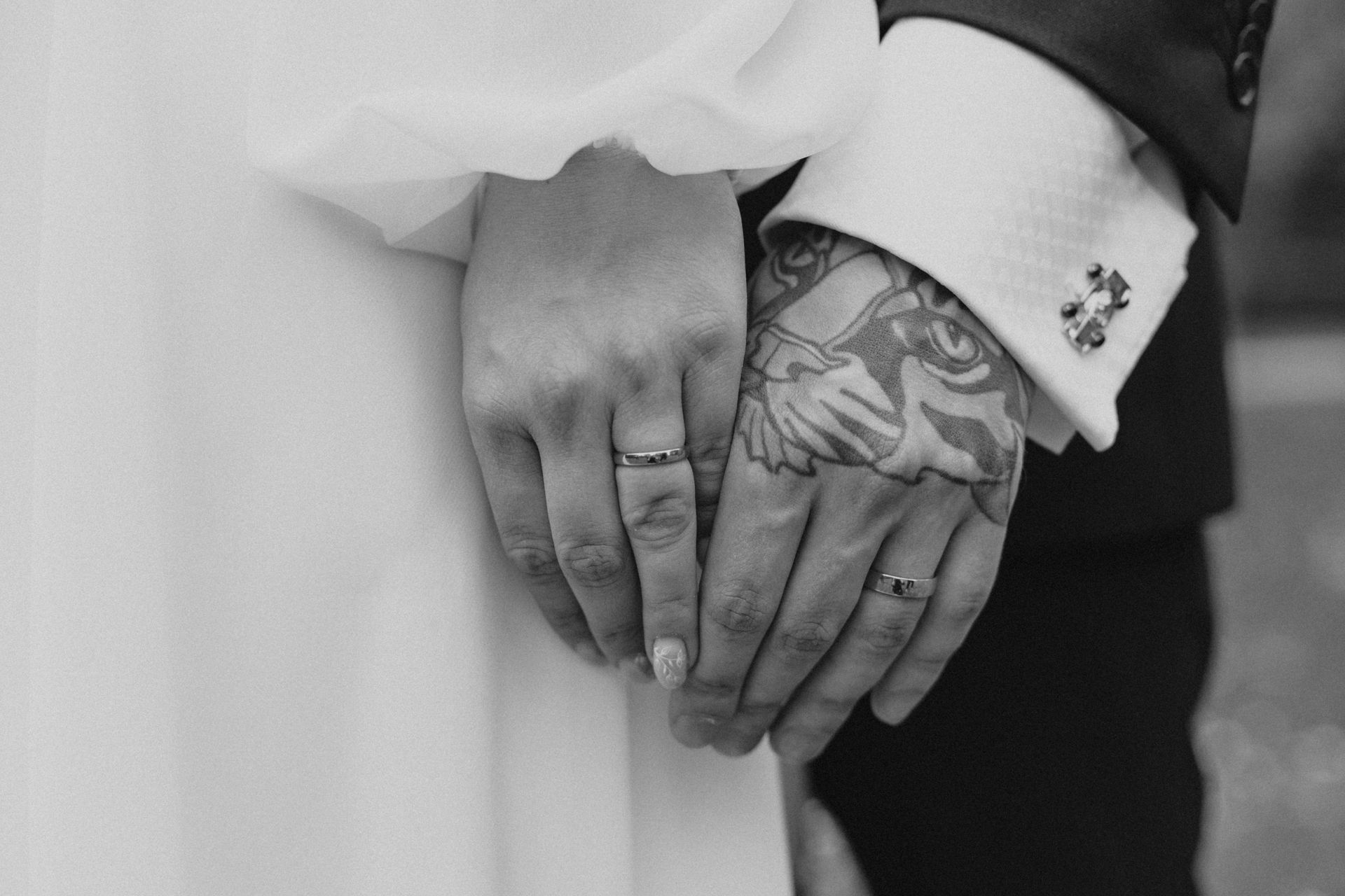 A close-up, black-and-white shot of a couple holding hands, highlighting wedding bands and a hand tattoo.