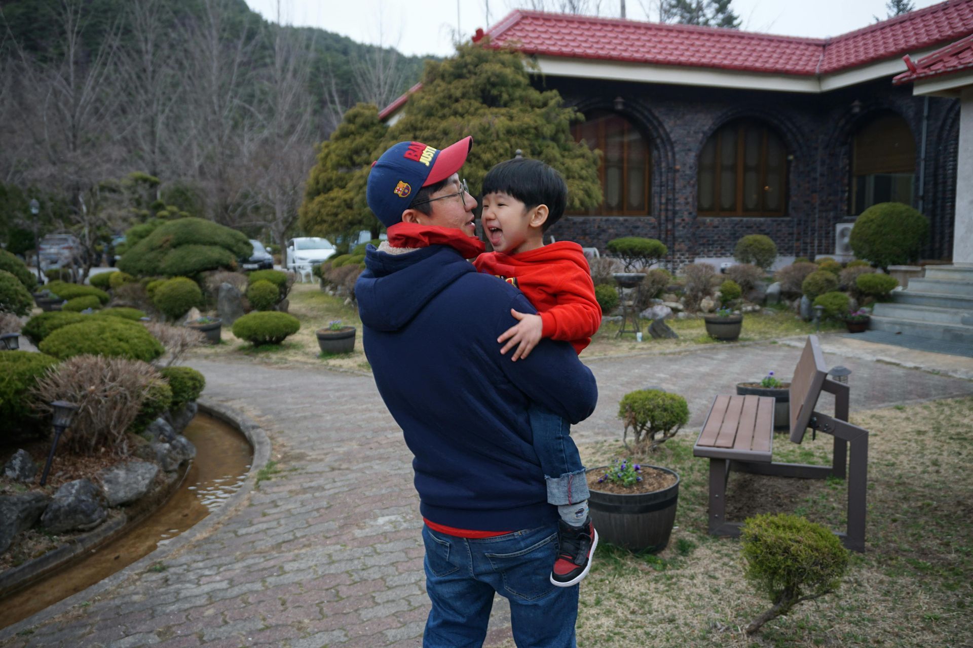 A person in a blue hoodie and cap holds a young child in a red jacket outdoors in front of a building with a red roof.