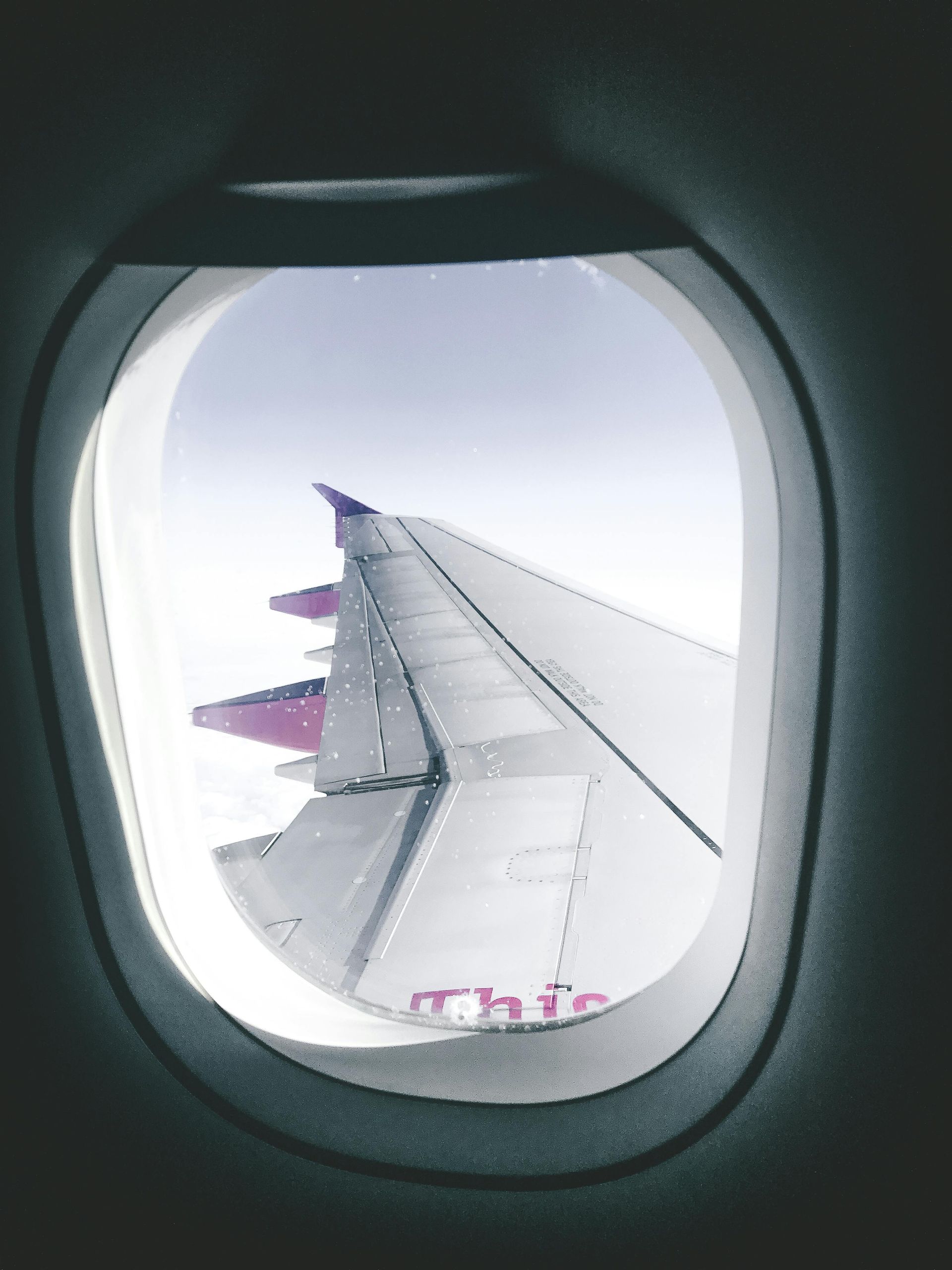 A view through an airplane window showing the wing of a Wizz Air plane against a bright, cloudy sky.
