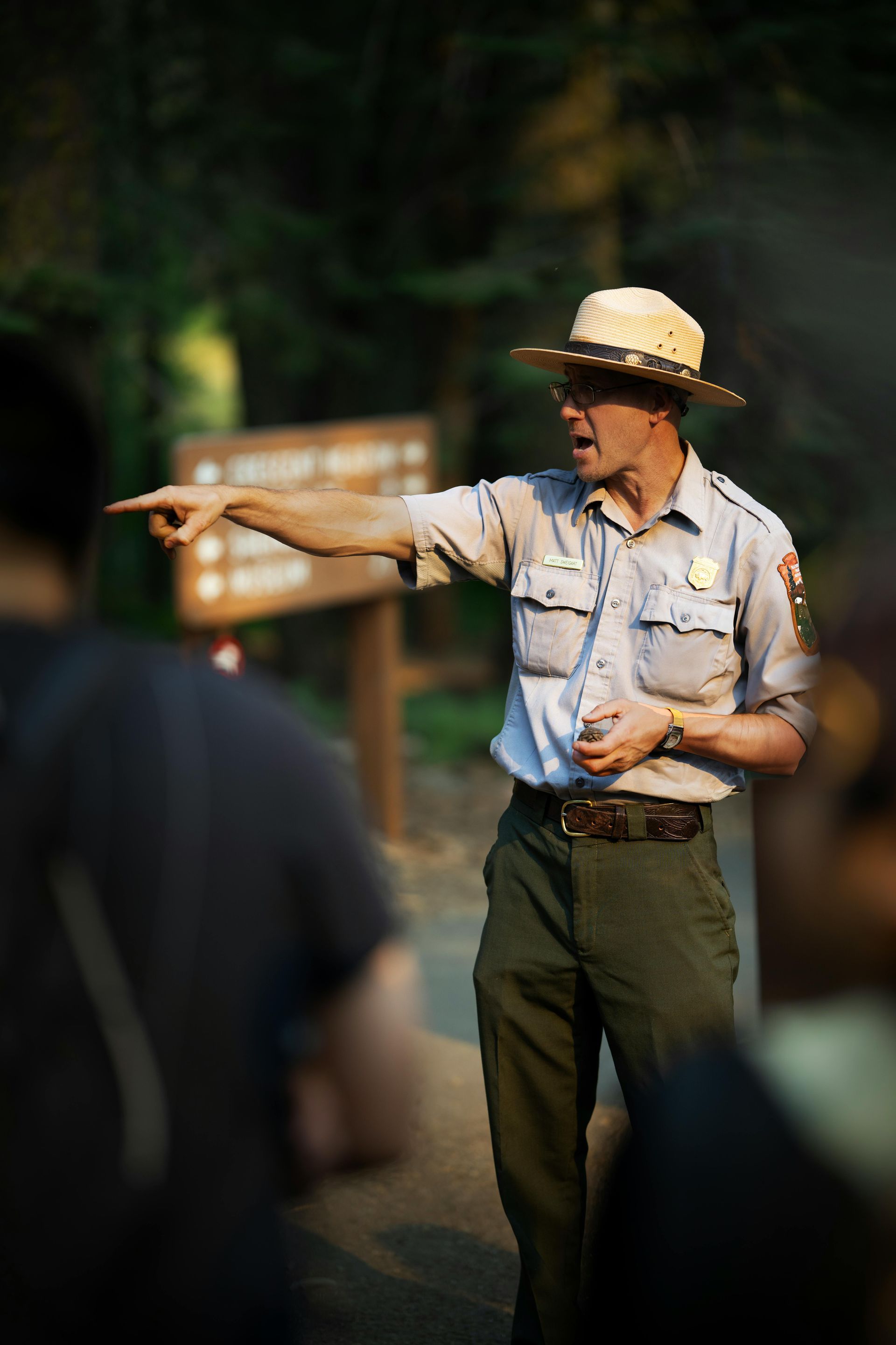 A park ranger in a tan uniform and hat points toward a trail sign in a forest.