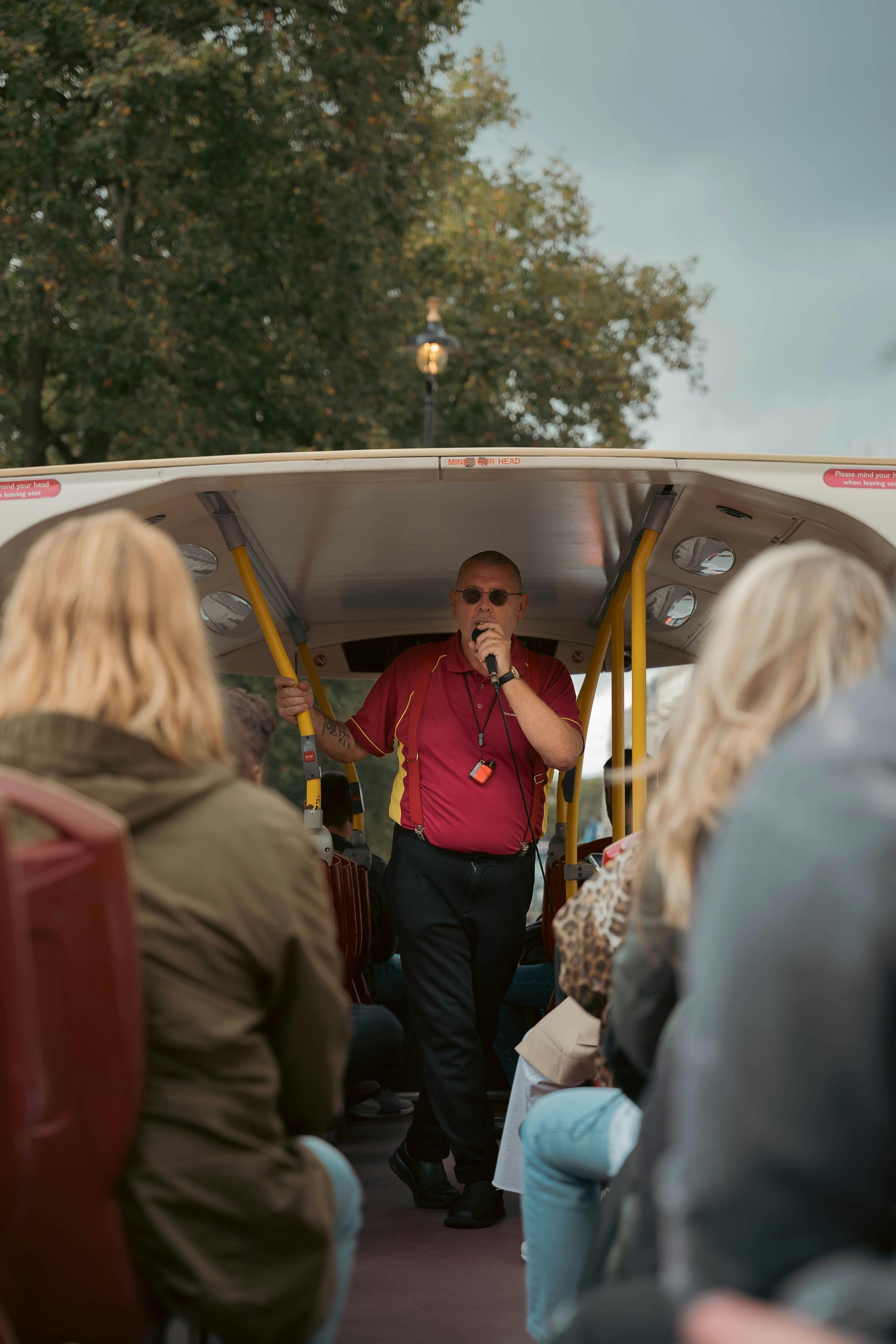 A tour guide in a red shirt speaks into a microphone while standing on an open-top bus, viewed from behind the passengers.