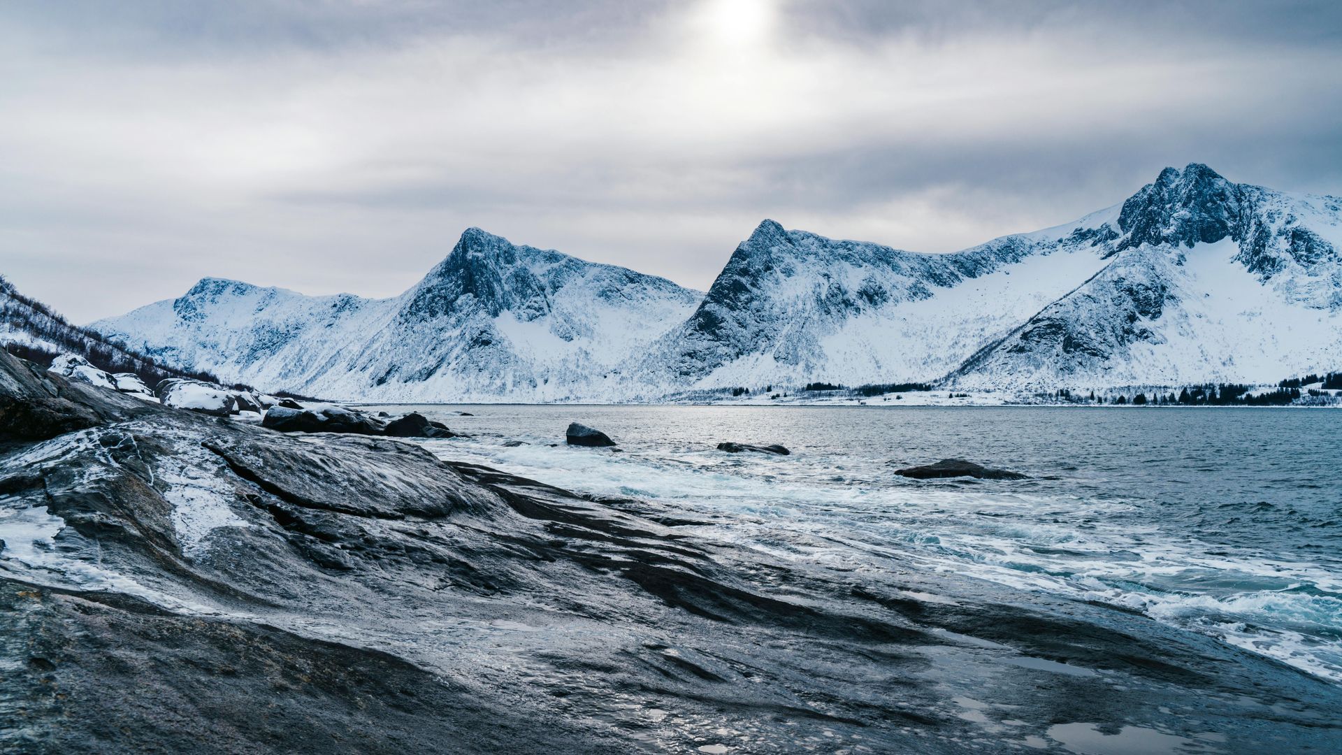 Snowy mountains rise above a rocky, ice-dusted coastline bordering a calm sea under a cloudy sky.