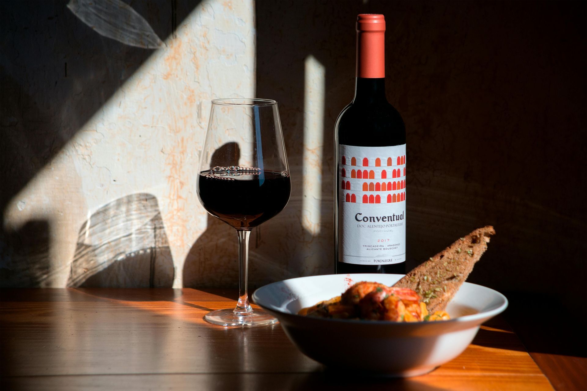 A bottle of red wine, a glass of wine, and a bowl of food on a wooden table, illuminated by a beam of sunlight.