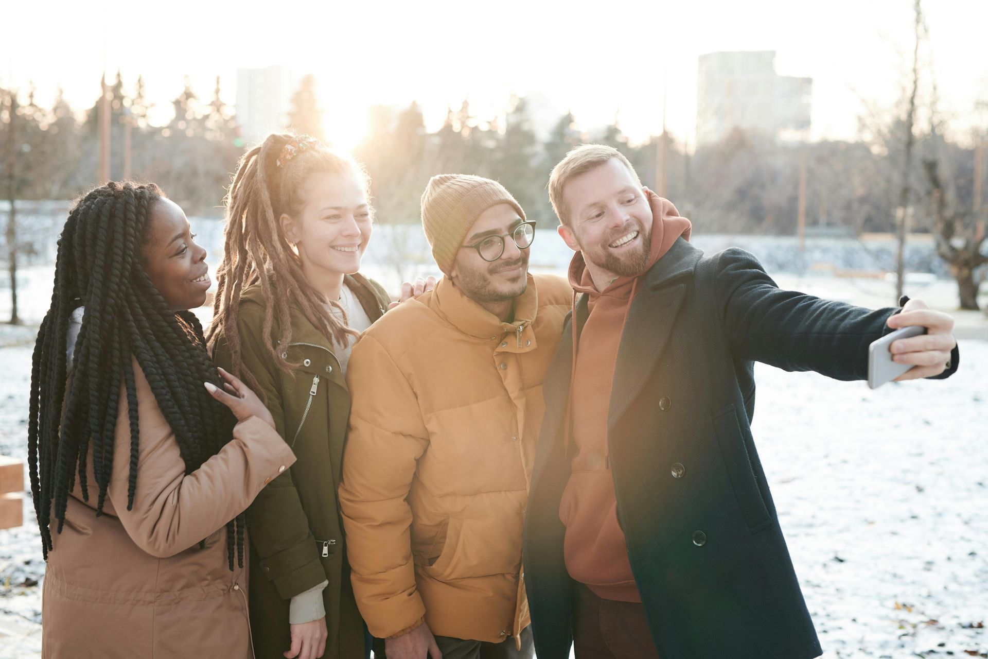 Four people smiling and taking a selfie outdoors in a snowy park during sunset.