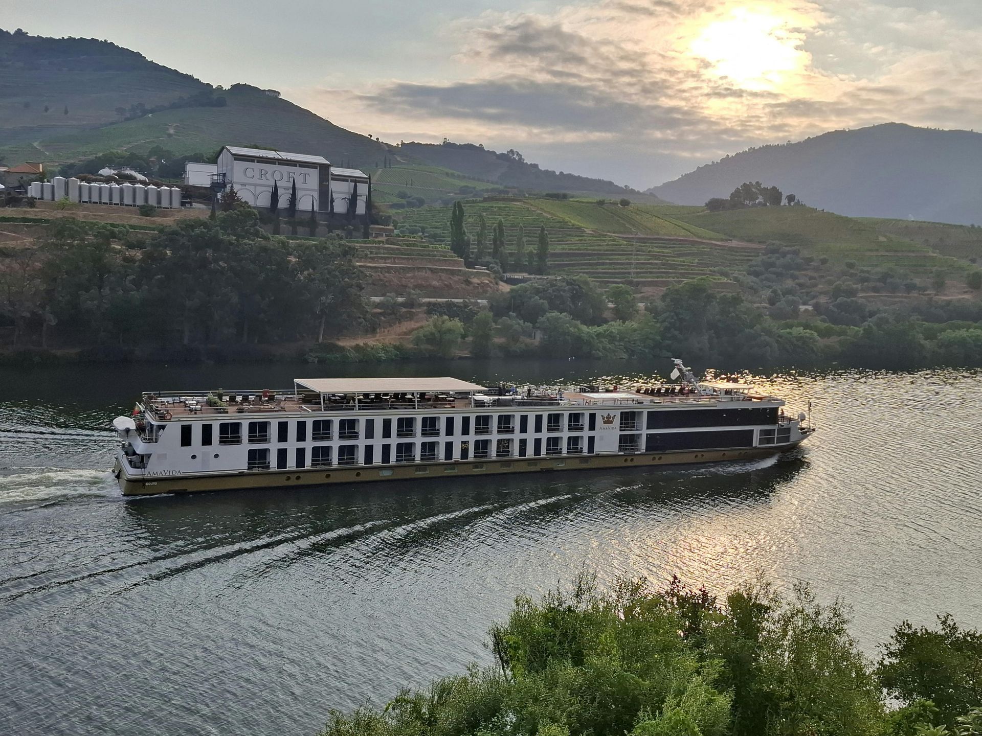 A long river cruise ship sailing on a wide river surrounded by terraced, hilly vineyards under a bright, sunny sky.